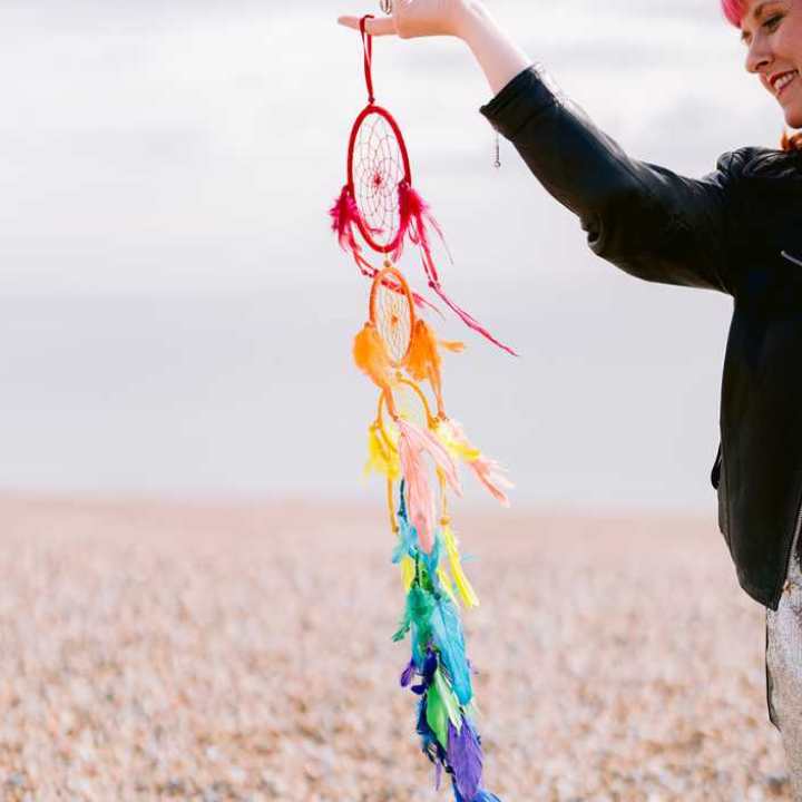 A rainbow coloured feather dream catcher being held in the wind on a beach.